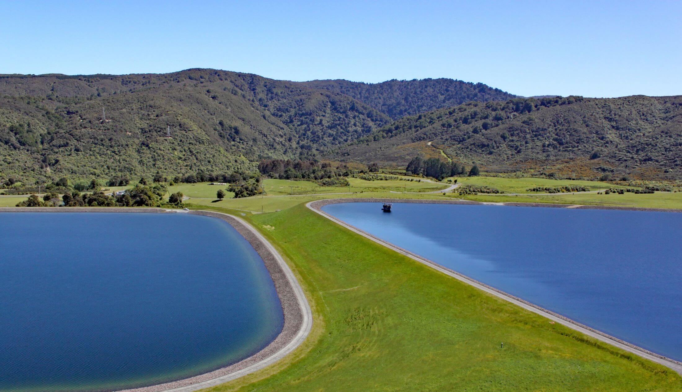 Aerial view of a reservoir with curved embankments, blue water basins, and surrounding green hills under a clear sky