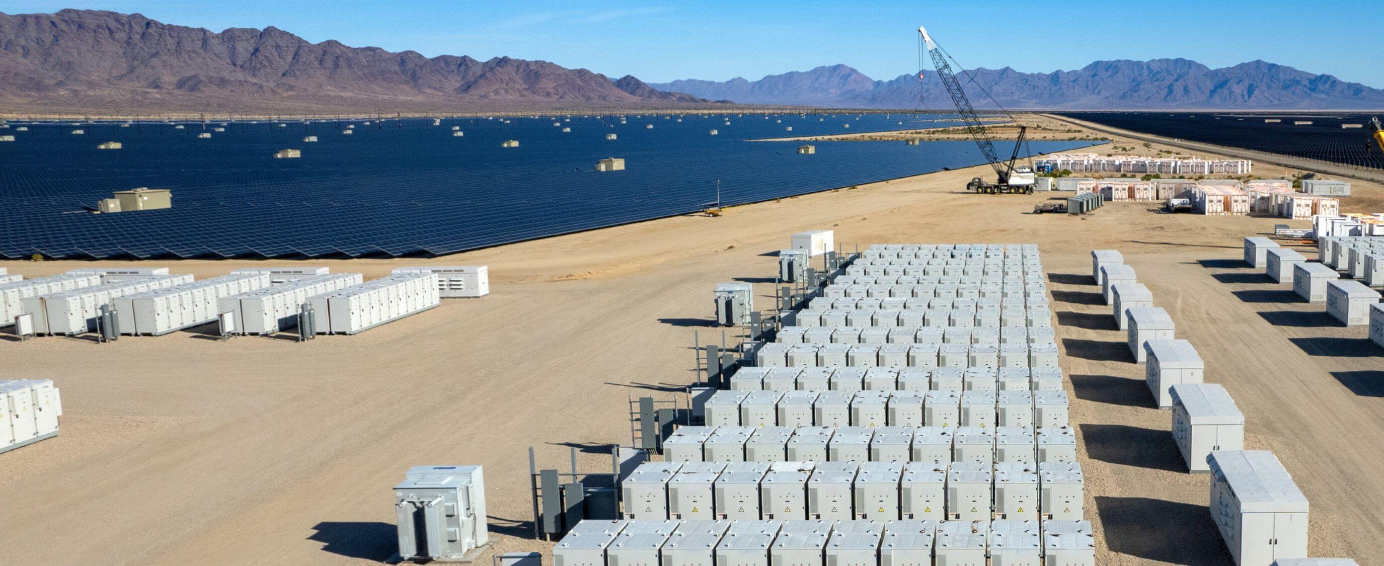 Utility-scale battery energy storage arrays installed beside a large solar power facility, with rows of equipment enclosures extending across a desert site and mountains visible in the distance.