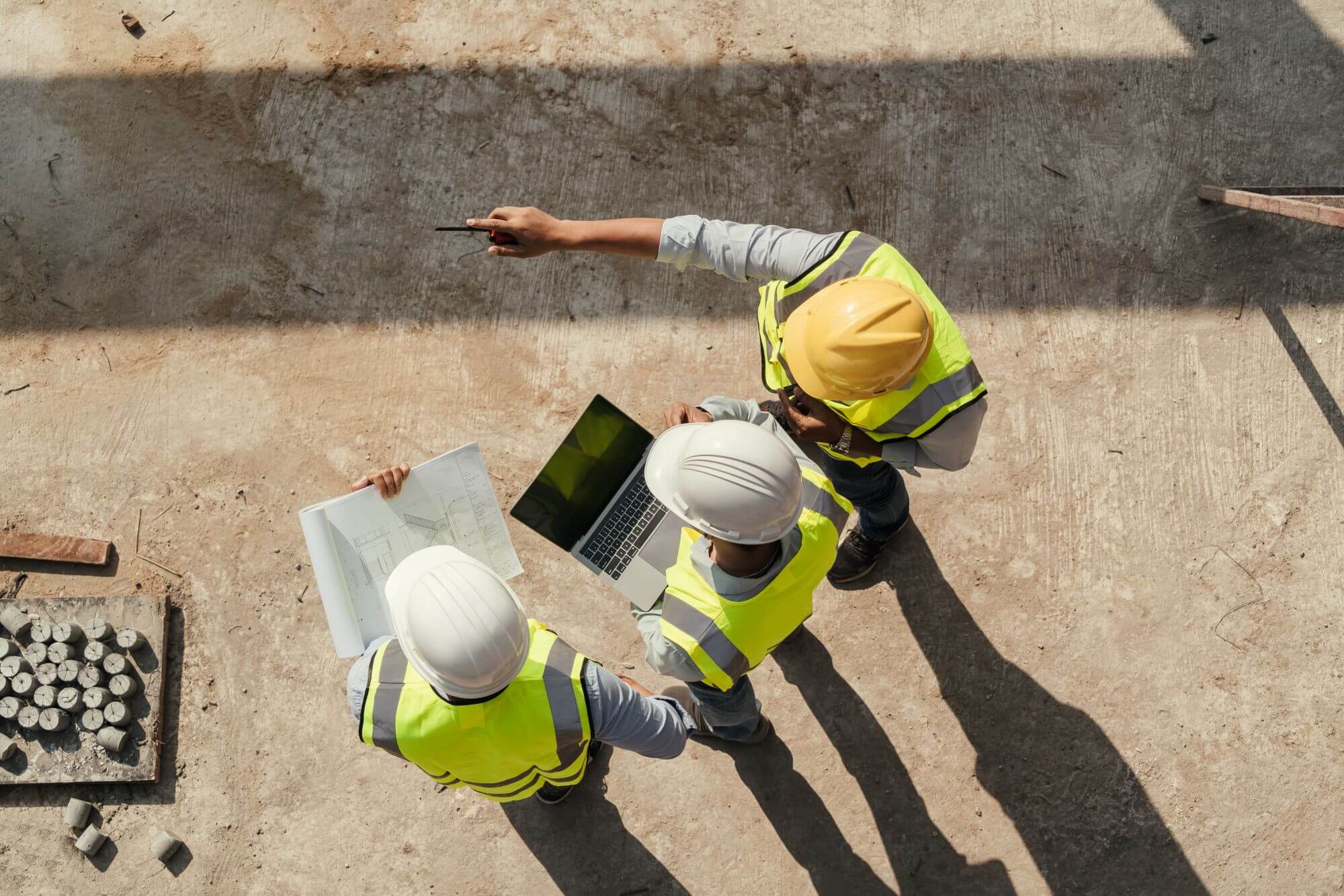 Overhead view of three engineering professionals in safety vests and hard hats reviewing blueprints and a laptop at a job site, with one pointing to a location on the ground.